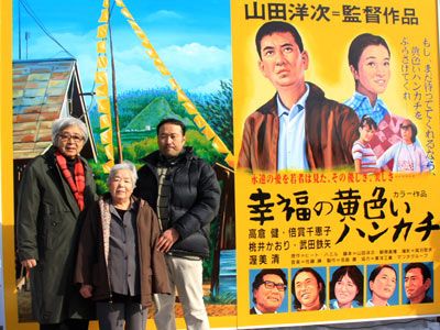 山田洋次と固い握手を交わす「幸福の黄色いハンカチ 想い出広場」管理人濱松フミさん－絵看板の前で